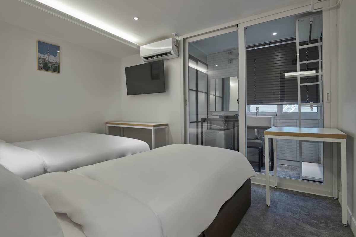Une chambre d’hôtel avec deux lits, une télévision, une fenêtre avec des stores et une petite table.