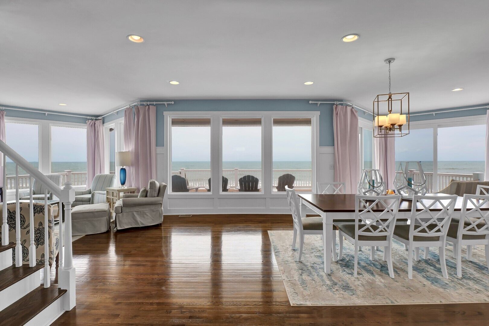 A spacious living room with a view of the ocean, a dining area with white chairs, and a staircase with white railings.