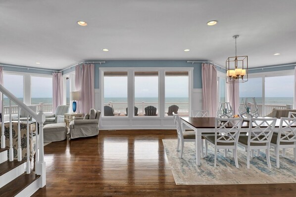 A spacious living room with a view of the ocean, a dining area with white chairs, and a staircase with white railings.