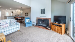 Una sala de estar con sofá, sillón, chimenea, televisión y una cocina al fondo.