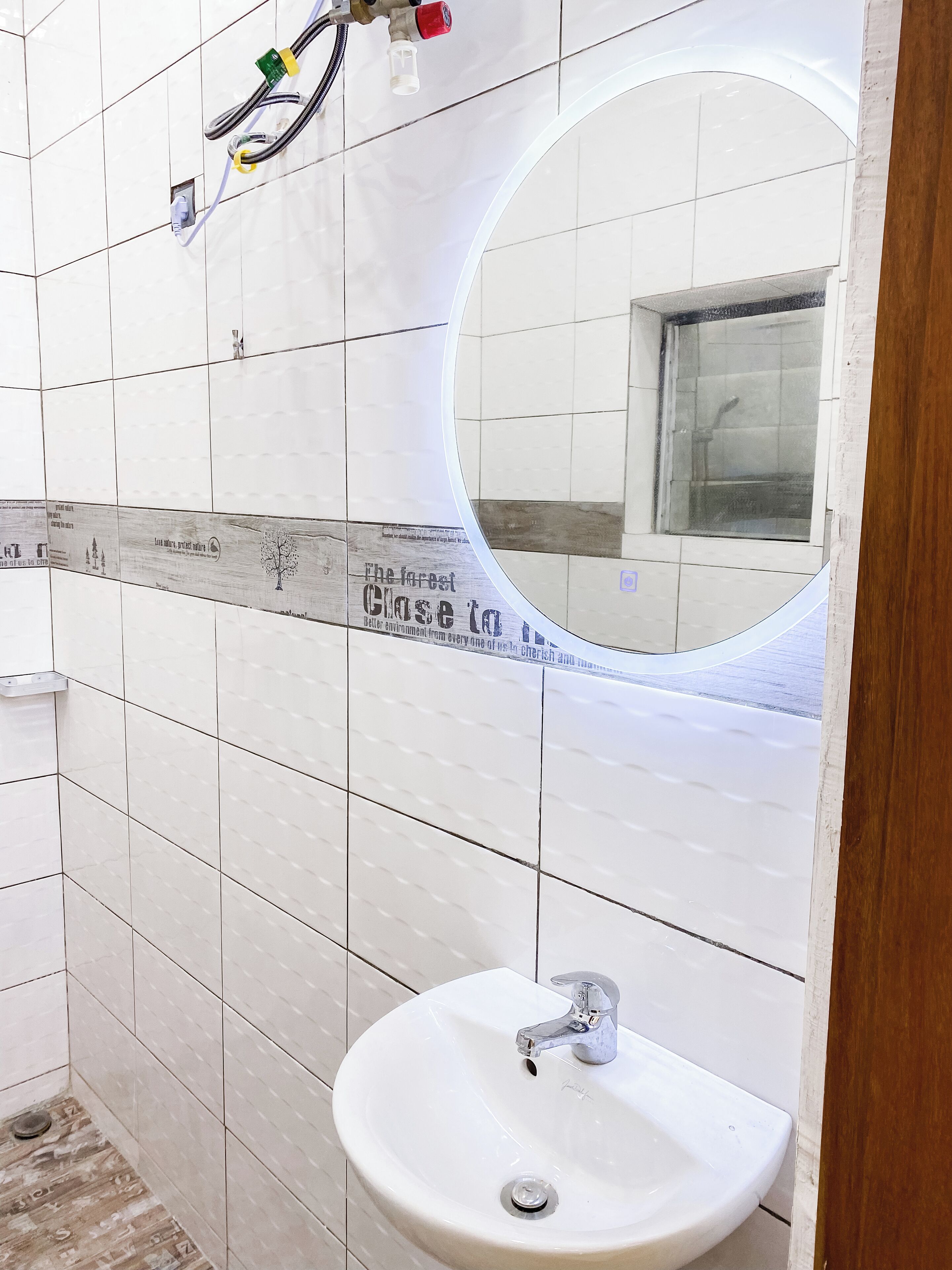 Ein Badezimmer mit weiß gefliesten Wänden, einem runden Spiegel, einem Waschbecken mit Armatur sowie sichtbaren Steckdosen und Kabeln.
