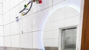 Ein Badezimmer mit weiß gefliesten Wänden, einem runden Spiegel, einem Waschbecken mit Armatur sowie sichtbaren Steckdosen und Kabeln.