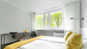 A hotel room with a bed, a desk, a chair, a window with curtains, and a view of trees outside. - MARGI Kai Apartament (Szczecin)