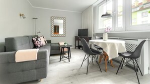 A modern living room with a grey sofa, a dining table with black chairs, a small side table, and a large window with curtains. - MARGI Kai Apartament (Szczecin)