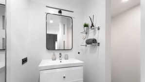 A modern bathroom with a white vanity, a mirror, and a shelf with toiletries. - MARGI Kai Apartament (Szczecin)