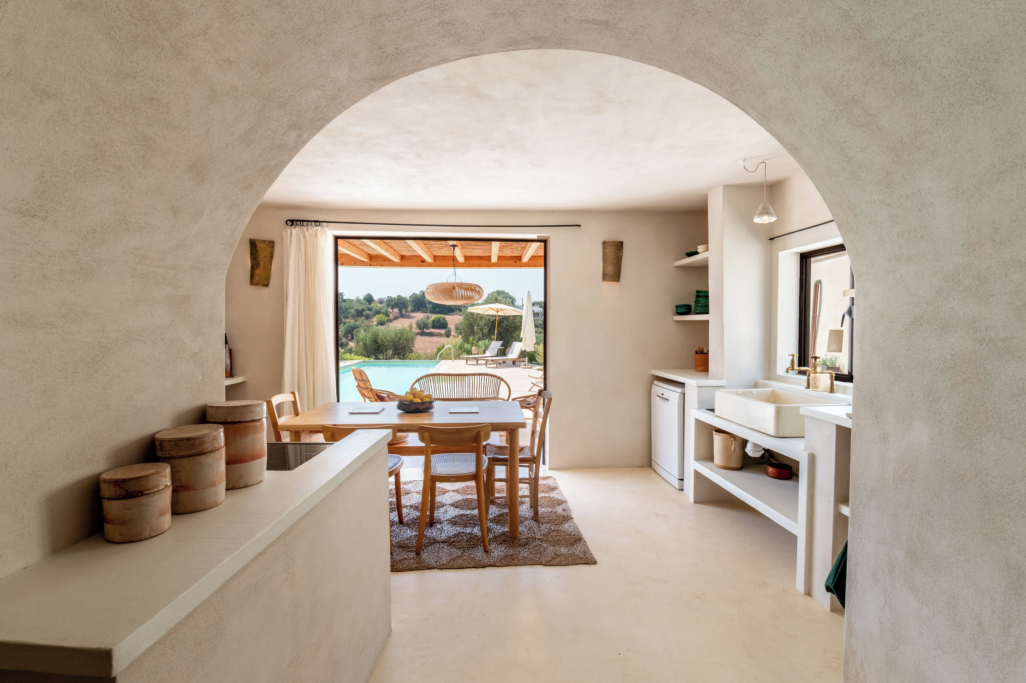A modern kitchen with an archway leading to a pool area, wooden furniture, and a view of the landscape.