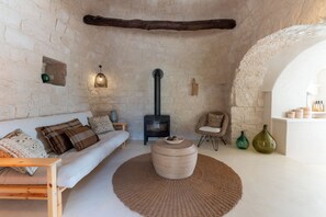 Um quarto aconchegante com parede de pedra, um sofá de madeira, um tapete redondo, um banquinho de fibra natural, uma cadeira de fibra natural, uma mesa de madeira, uma luminária na parede, uma lareira a lenha preta e uma estante com potes.