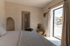 Quarto com cama, janela com cortinas, um banquinho de madeira e uma garrafa verde na parede.
