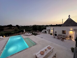 Área de piscina com espreguiçadeiras e uma mesa de jantar, cercada por um edifício branco com telhado de pedra.