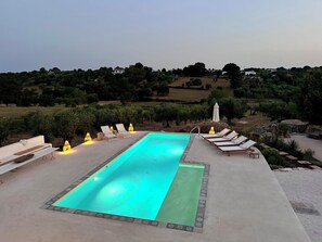 Uma piscina retangular com um tapete verde no centro, rodeada por cadeiras de lounge brancas e pequenas mesas.
