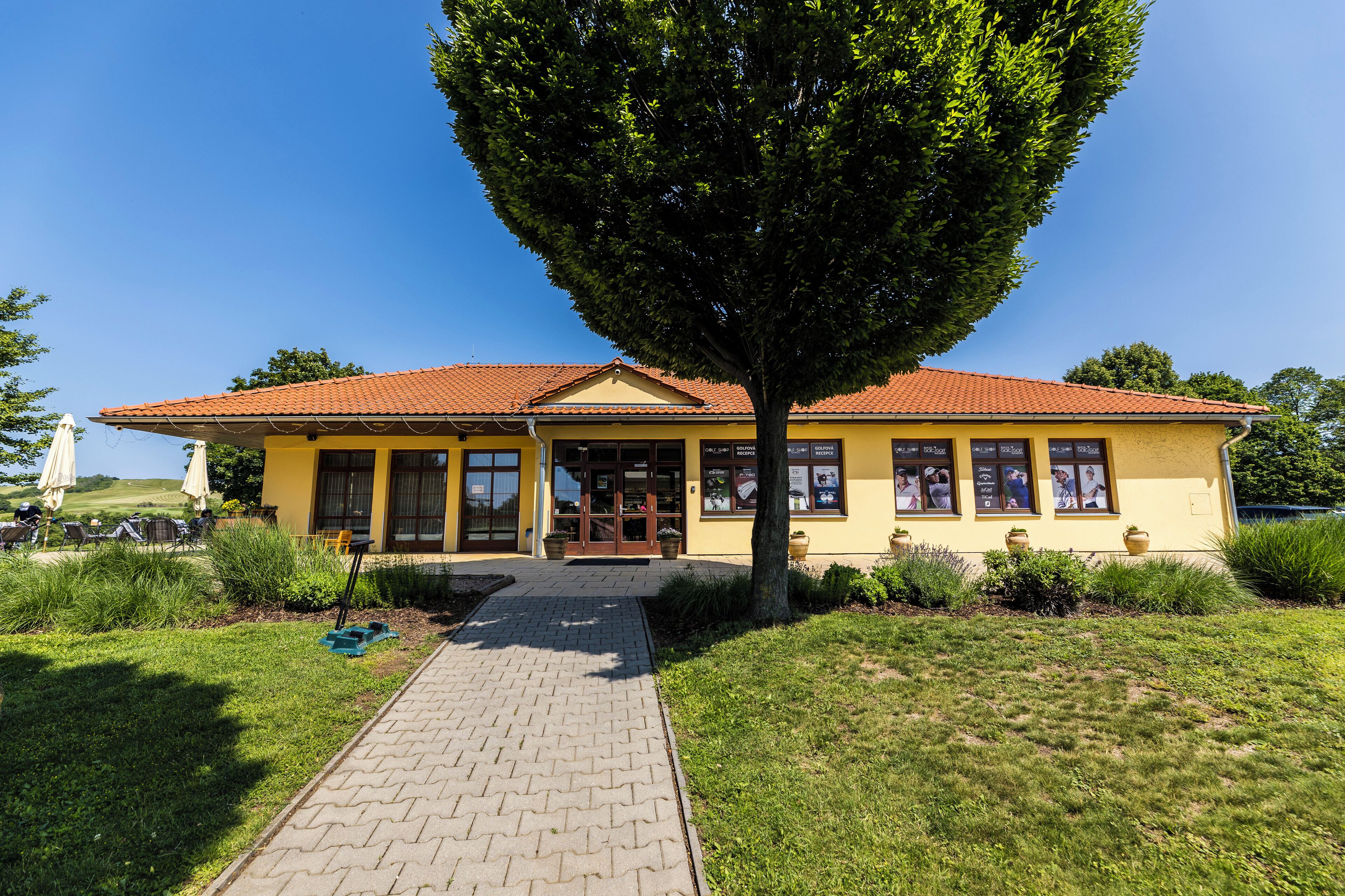 Un edificio giallo con tetto in tegole rosse, ingresso lastricato e un grande albero di fronte.