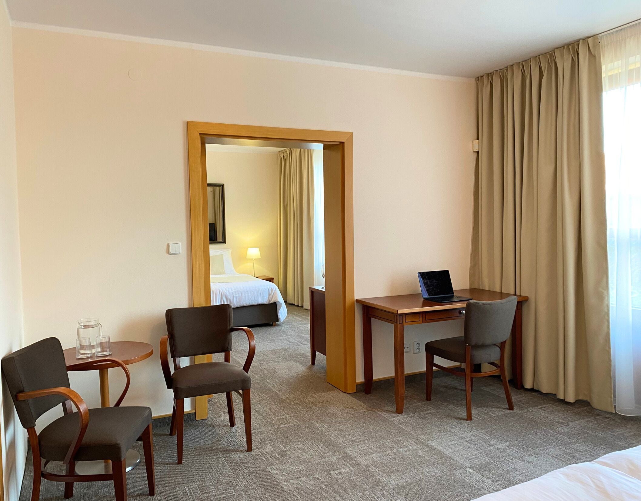 A hotel room with a bed, a desk with a laptop, two chairs, a small table with glasses, and a mirror.