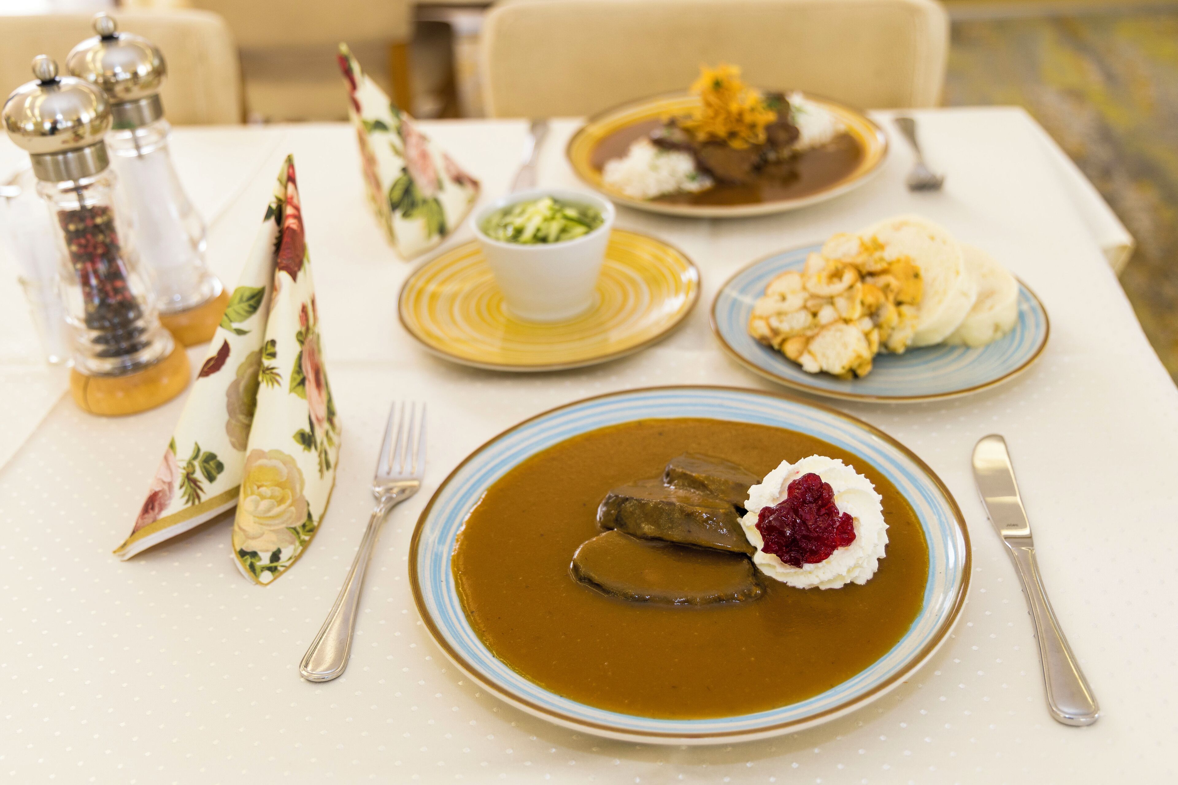 Una mesa dispuesta con platos de comida, incluyendo un guiso con una cucharada de crema y una porción de carne, además de un bol de verduras verdes.
