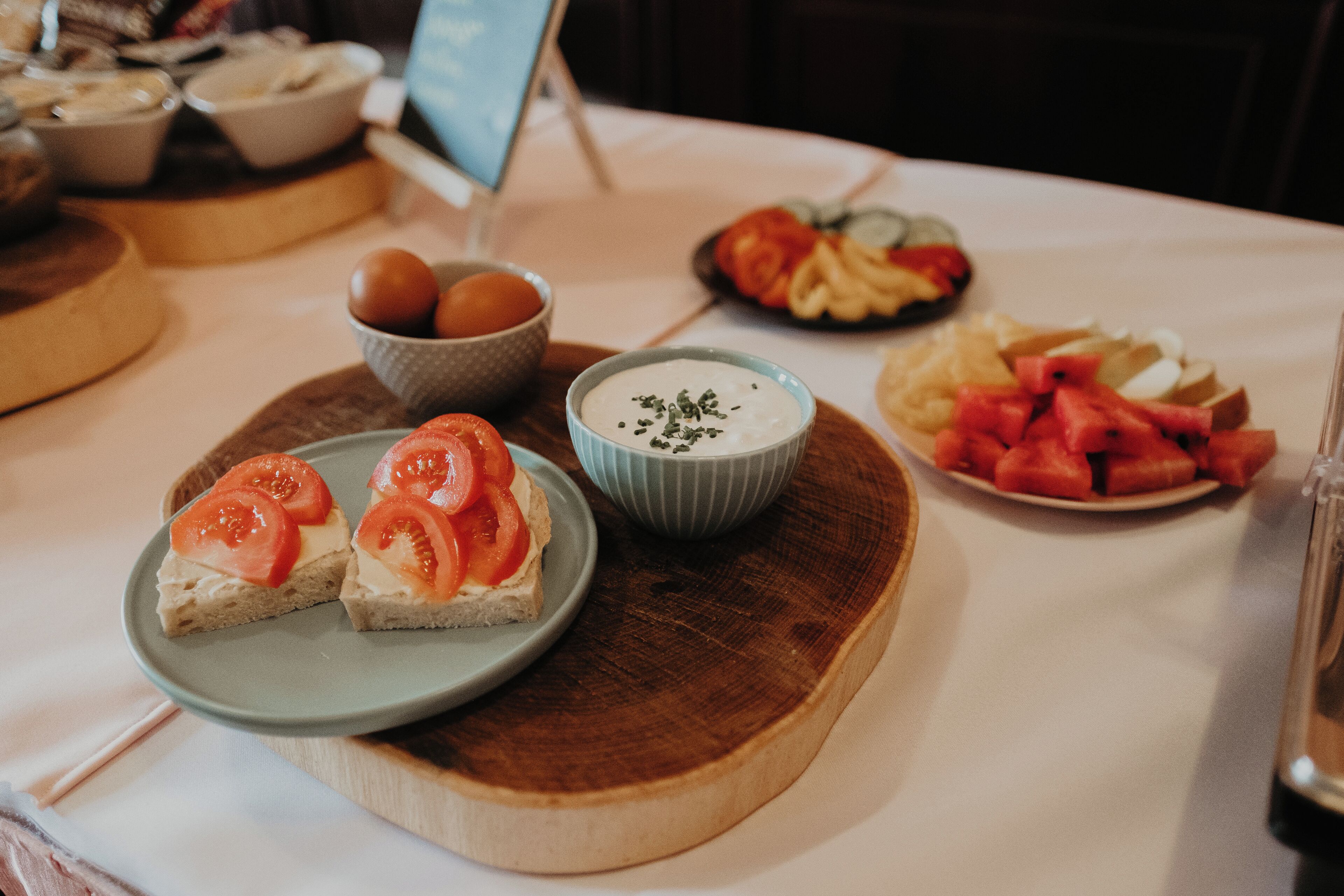 Una mesa con diversos alimentos, incluyendo pan con rodajas de tomate, un bol de salsa, y platos con frutas y verduras cortadas.