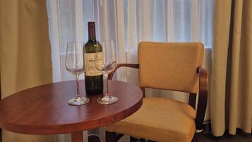 Une table ronde en bois, une bouteille de vin et deux verres, une chaise en osier et une vue sur les arbres à travers de fines rideaux.