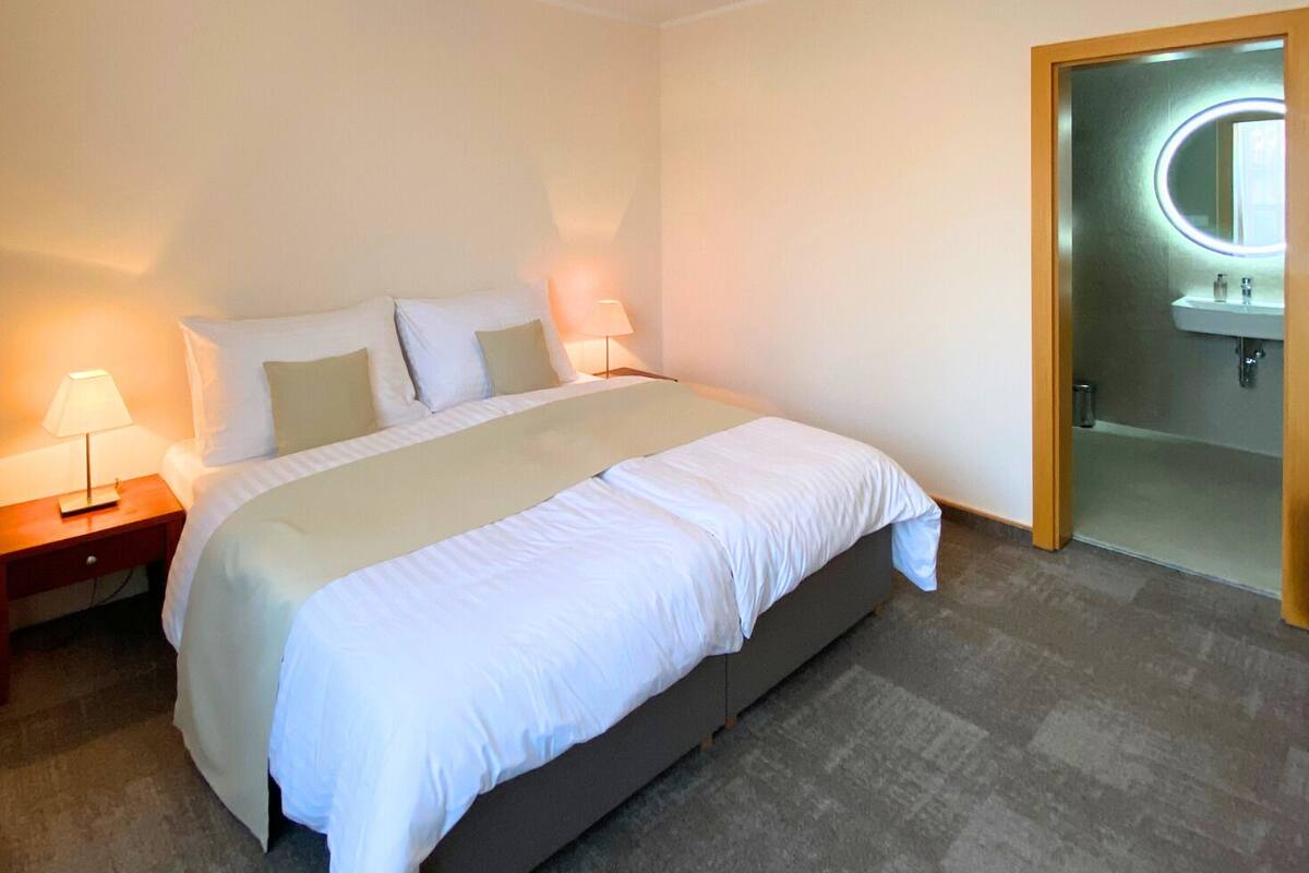 Une chambre d’hôtel bien rangée, avec un lit, des lampes de chevet et une salle de bain visible à travers une porte ouverte.