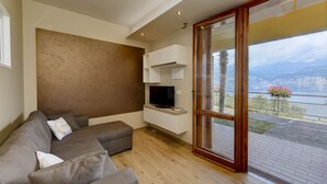 A modern living room with a grey sofa, a flat-screen TV, and a large sliding glass door leading to a balcony with a city view.