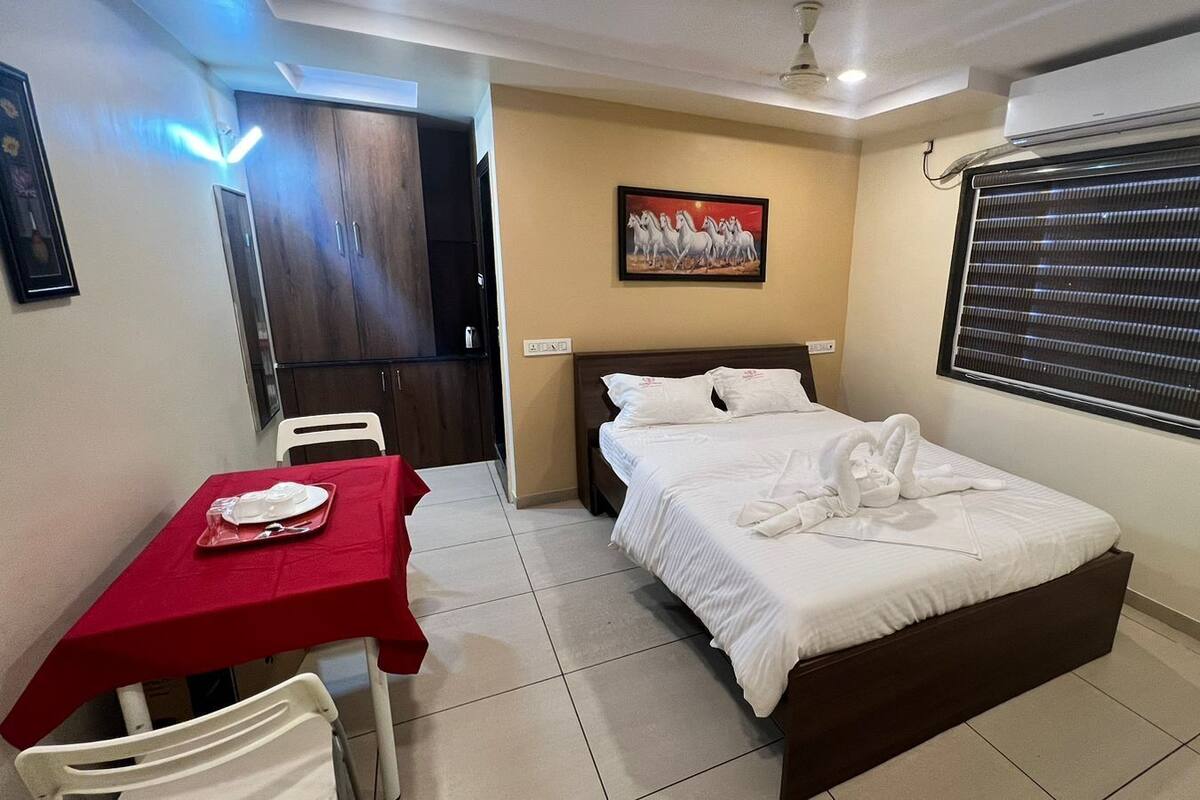Una habitación de hotel con una cama, una mesa de comedor con mantel rojo, un cuadro en la pared y una ventana con persianas.