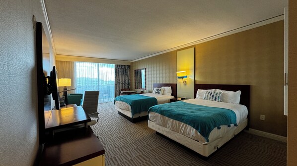 A hotel room with two beds, a desk, a chair, a TV, and a large window.