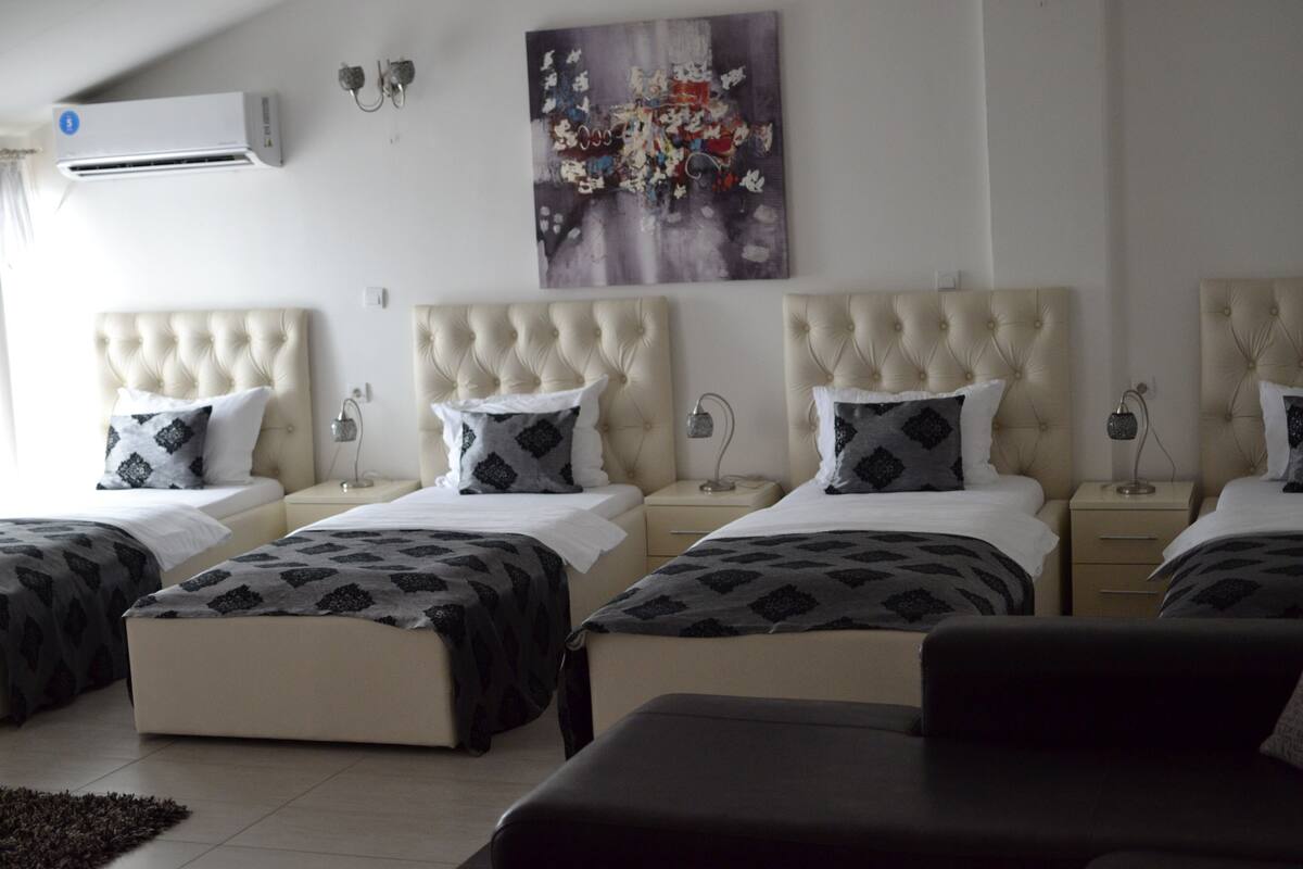 Ein Hotelzimmer mit vier nebeneinander stehenden Betten, jedes mit einer gepolsterten Kopfstütze und gemusterten Bettwäsche. Darüber an der Wand hängt ein großes abstraktes Gemälde.