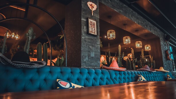 Una zona de bar con un banco acolchado azul, decoraciones de cactus y lámparas colgantes.