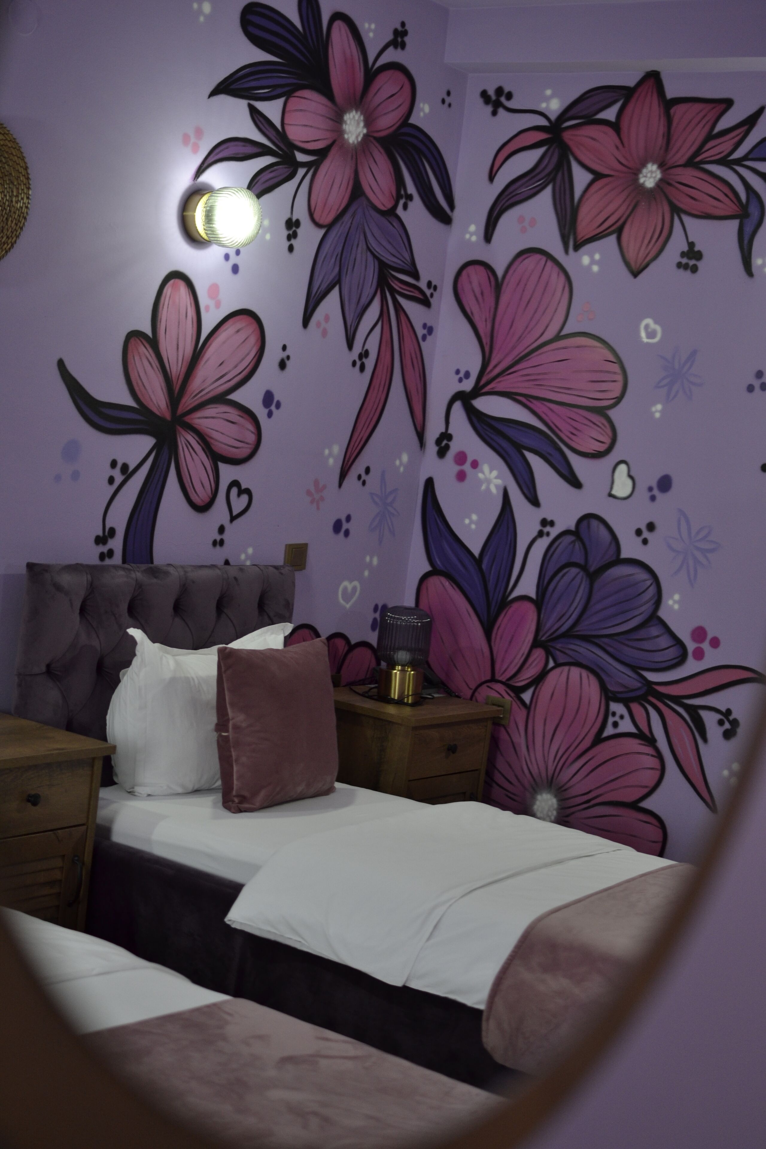 Un dormitorio con cabecera capitonada, cama con ropa de cama blanca y rosa, mesita de noche con lámpara y una pared decorada con motivos florales.