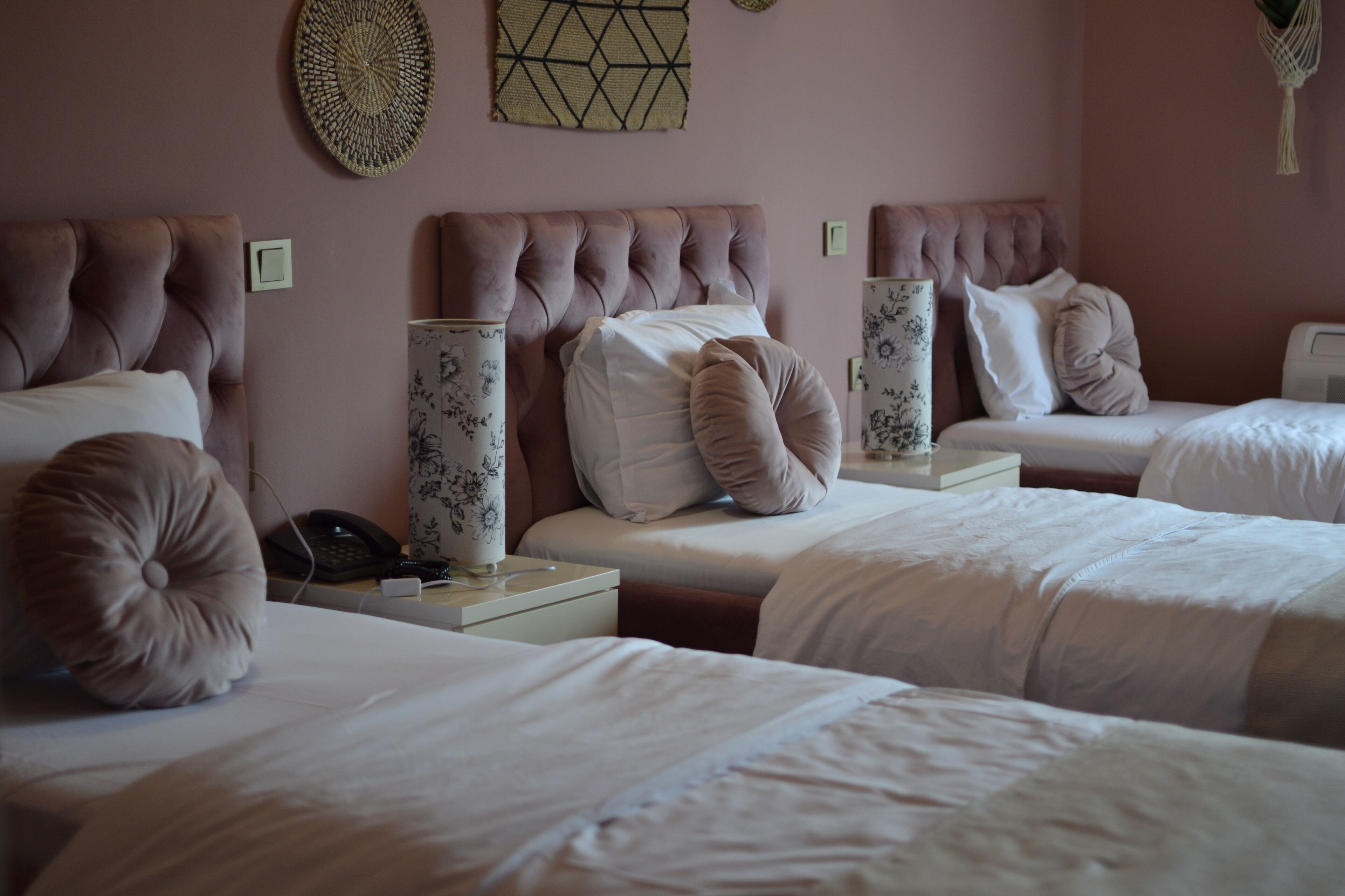 A hotel room with three beds, each with a tufted headboard and white bedding. There are decorative wall hangings above the beds and a bedside table with a lamp and a phone.