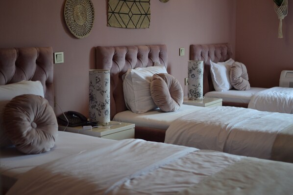 A hotel room with three beds, each with a tufted headboard and white bedding. There are decorative wall hangings above the beds and a bedside table with a lamp and a phone.