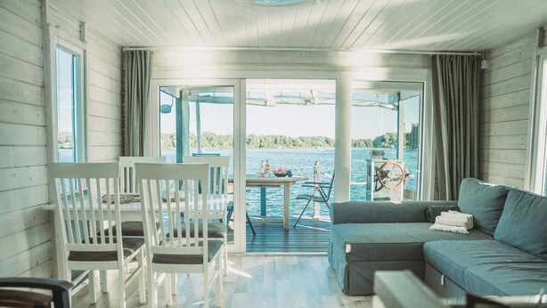 Ein gemütliches Zimmer mit Holzboden, einer grauen Couch, einem Esstisch mit Stühlen und Seeblick durch große Fenster.