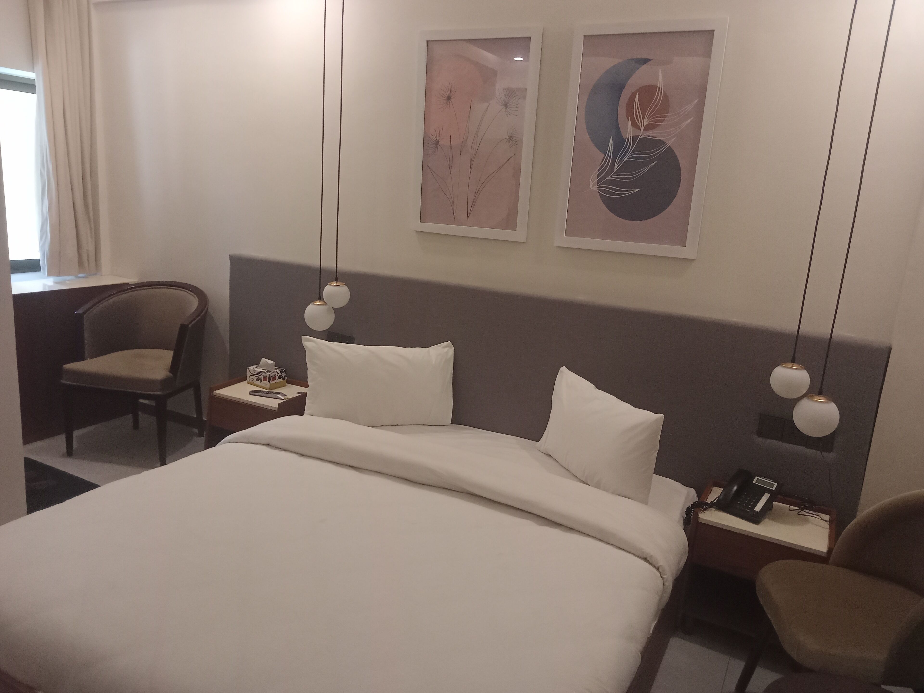Een hotelkamer met een bed, twee stoelen, een nachtkastje met een telefoon, en twee kunstwerken aan de muur.