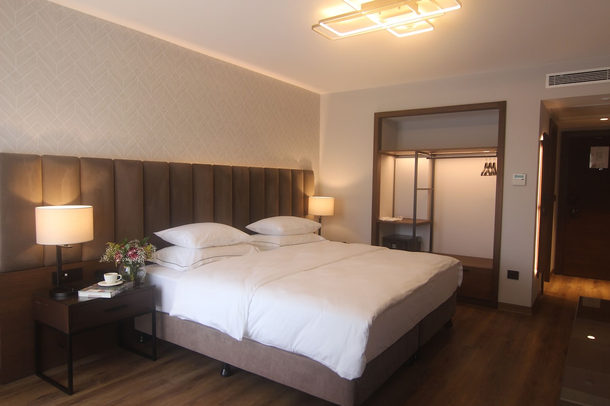 A hotel room with a large bed, two bedside tables with lamps, a nightstand with a cup and book, and a mirror on the wall.