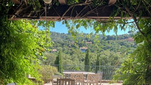 Una veranda esterna con pavimento in pietra e mobili in legno, tra cui un tavolo e sedie, sotto una pergola con vista su un paesaggio verde e edifici in lontananza.