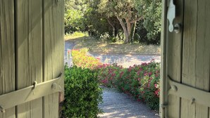 Una porta in legno aperta che dà su un giardino con un vialetto, circondato da vegetazione e piante fiorite.