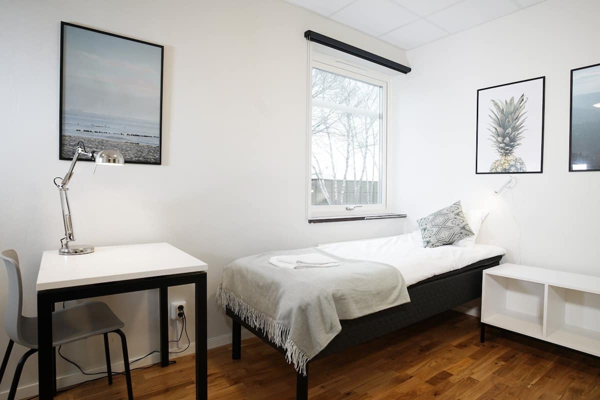 A small, modern room with a bed, a desk, and a chair. There are two framed pictures on the wall, one of a landscape and one of a pineapple.