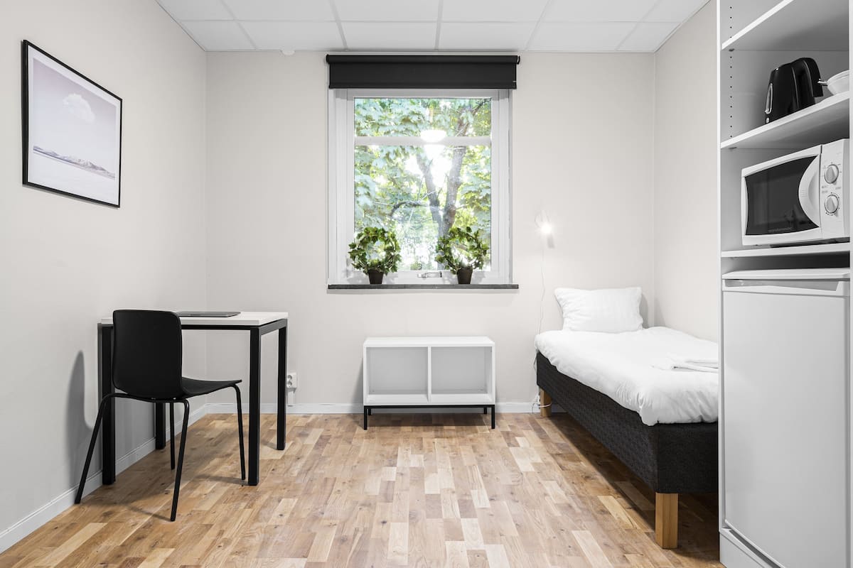A modern, minimalist room with a wooden floor, a black chair, a white desk, a bed with white bedding, a white bench, a window with a view of trees, and a microwave on the right.
