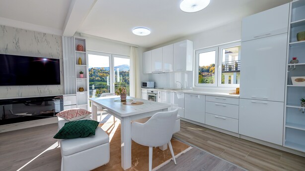 A modern kitchen with white cabinetry, a dining area with a white table and chairs, a flat-screen TV, and a view of a balcony with a mountain backdrop. - Green Hills Resort (Hadzici)