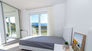 A modern bedroom with a large bed, a small bedside table, a window with a view of mountains, and a sliding glass door leading to an outdoor area. - Green Hills Resort (Hadzici)