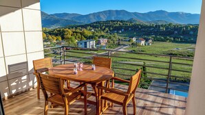 A balcony with a wooden table and chairs, overlooking a scenic view of mountains and a village. - Green Hills Resort (Hadzici)