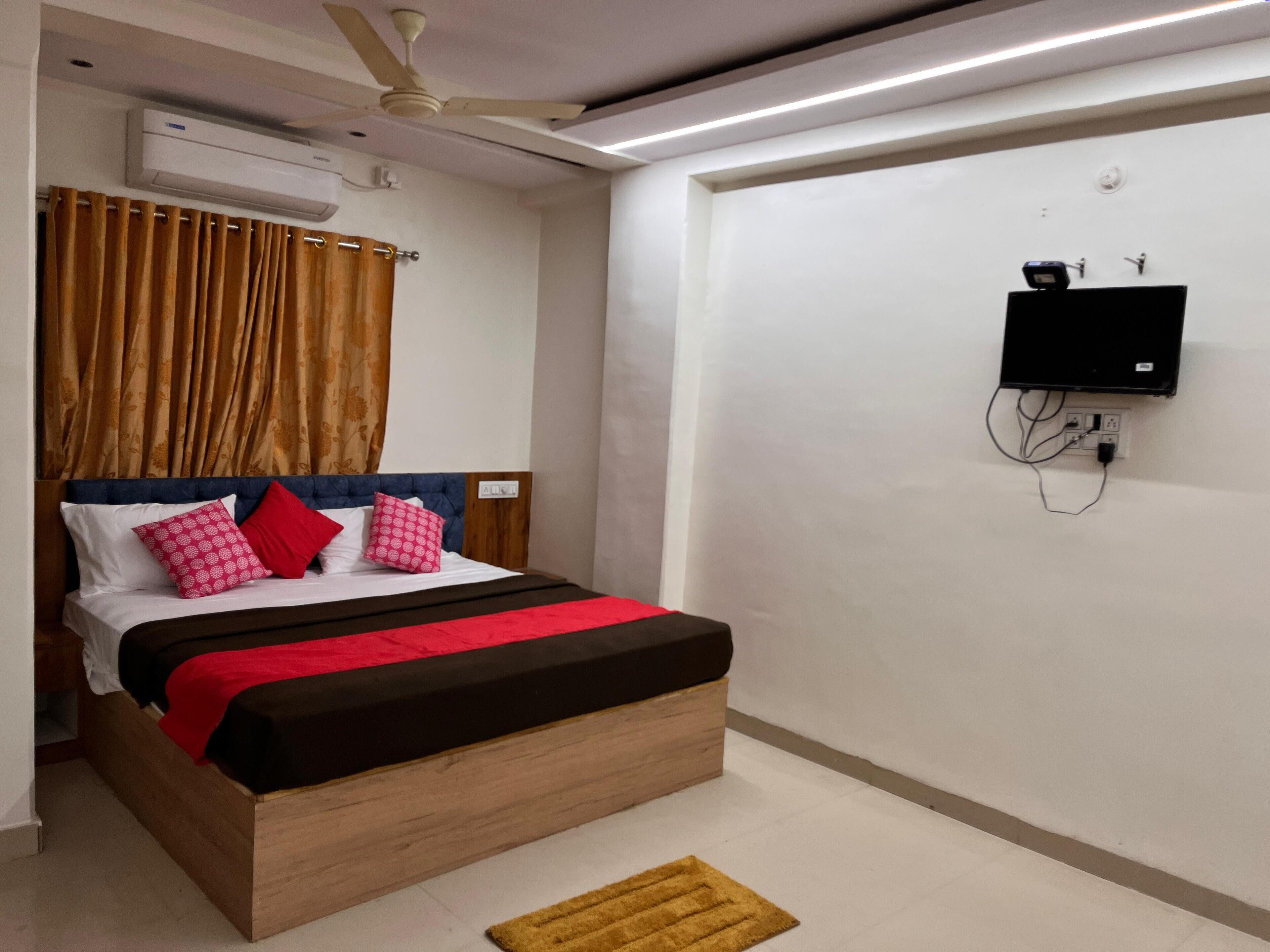 A bedroom with a bed, curtains, a ceiling fan, an air conditioning unit, a TV mounted on the wall, and a power outlet.