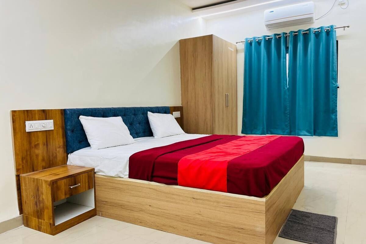Een slaapkamer met een houten bedframe, witte beddengoed, een rode deken, een nachtkastje, een houten kledingkast en een raam met turquoise gordijnen.