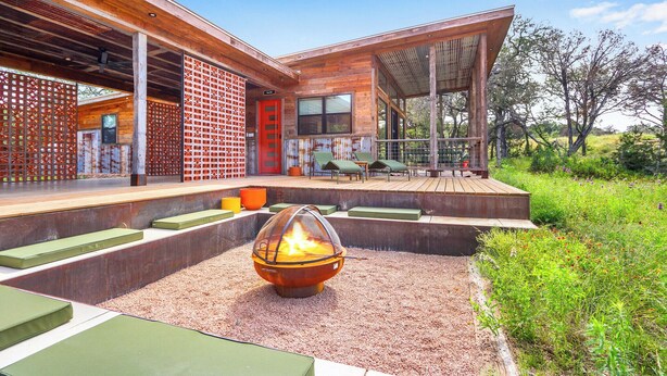 Ein modernes Haus mit Feuerstelle in der Mitte, Sitzgelegenheiten im Freien und einer überdachten Terrasse.