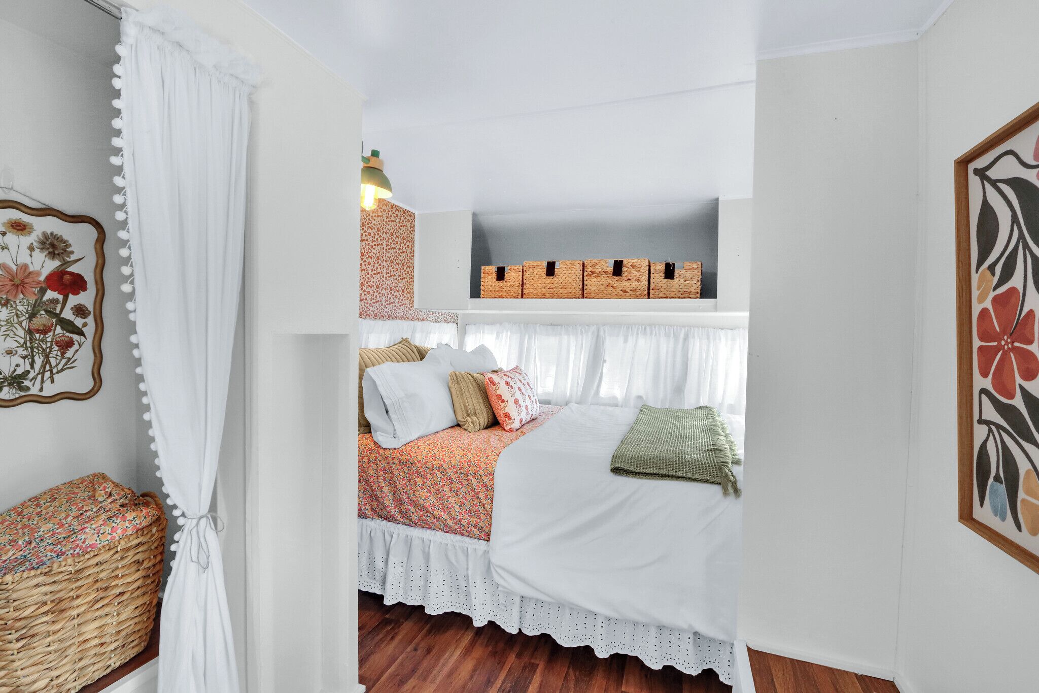 Ein gemütliches Schlafzimmer mit Bett, einer Ablage mit Aufbewahrungsboxen, einem Korbwäscher und einem floralen Bild in einem Rahmen an der Wand.