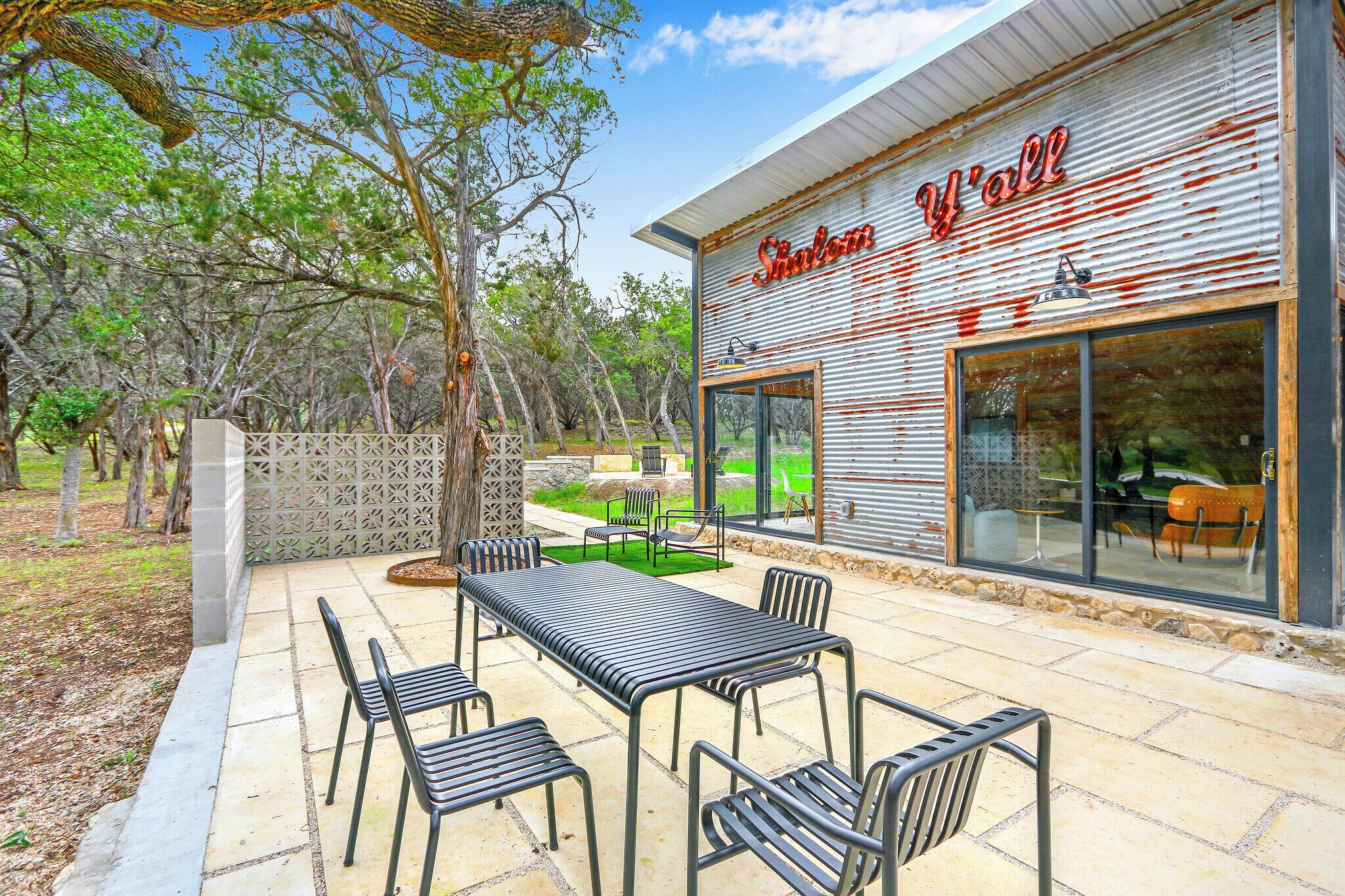 A modern outdoor patio area with a metal table and chairs, a glass door entrance, and a view of the surrounding trees.