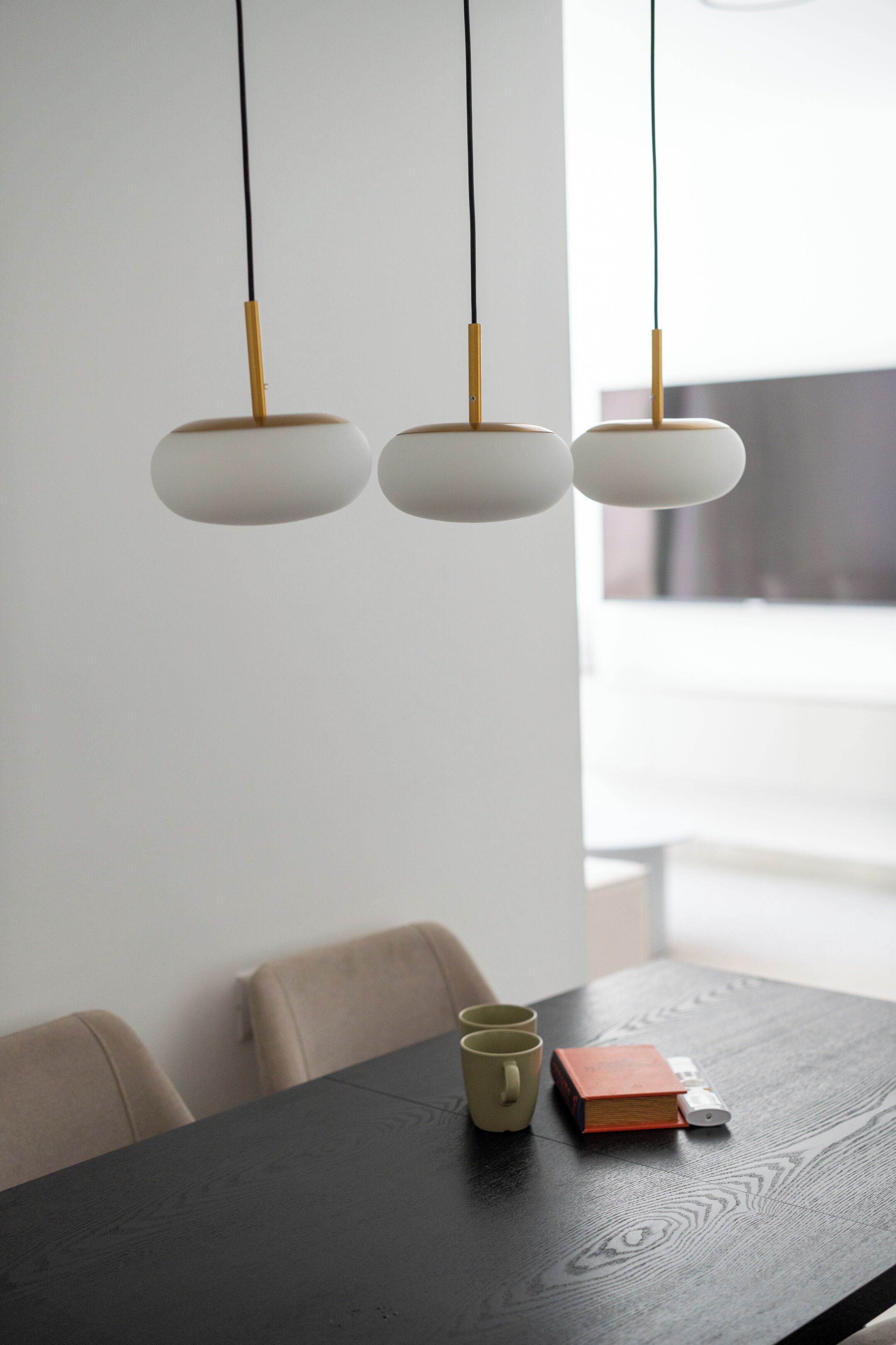Une table à manger en bois foncé, trois suspensions au design minimaliste, ainsi qu’un petit livre et deux tasses posés sur la table.