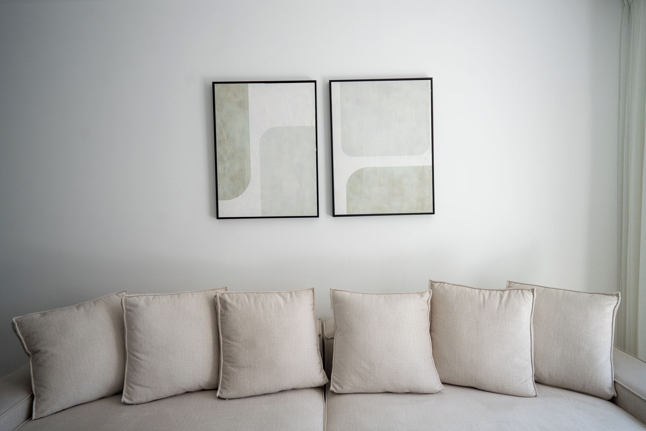 Un sofá en tonos neutros con seis cojines, una pared lisa y dos obras de arte abstracto.
