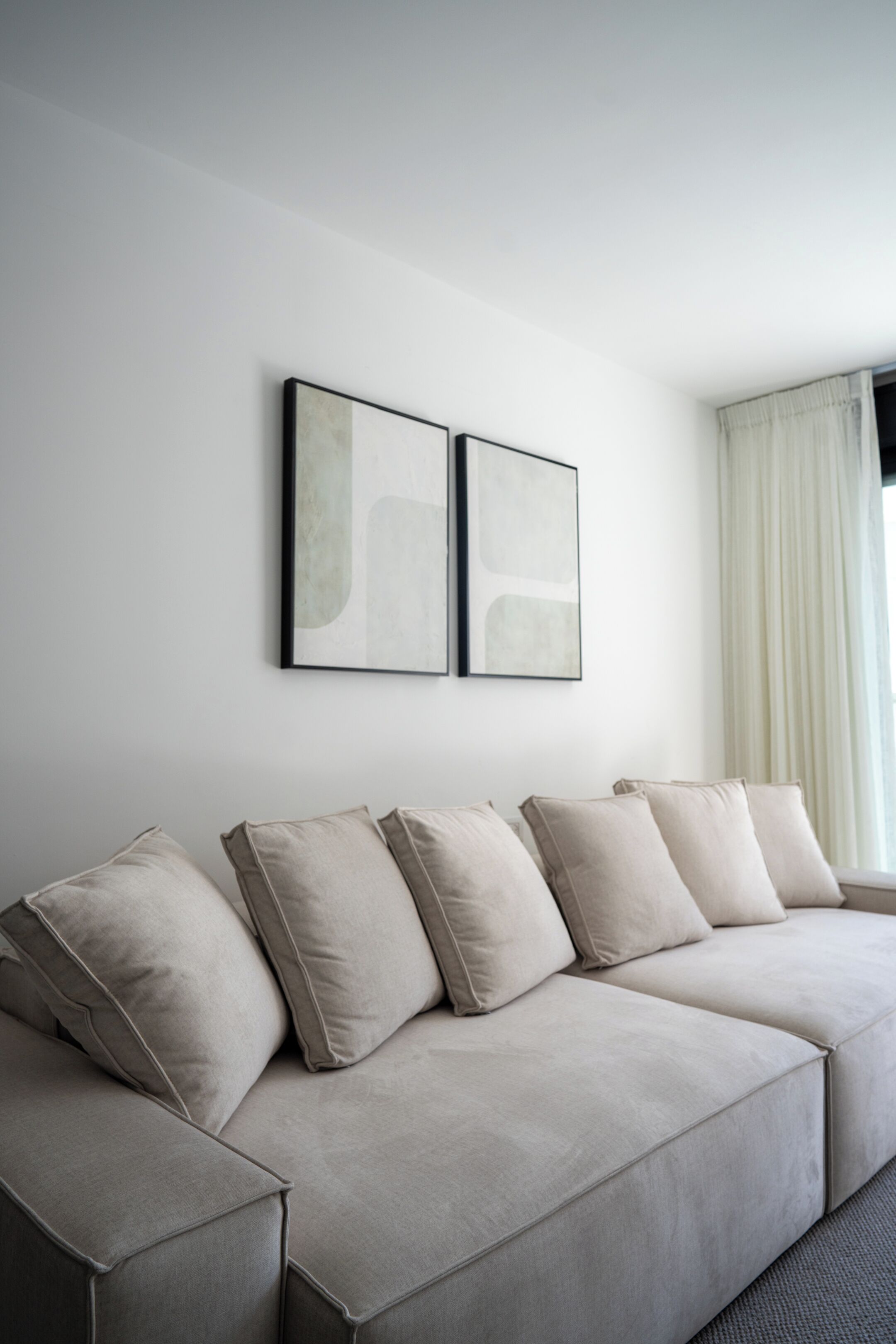 Una sala de estar moderna con un sofá seccional beige, dos cuadros abstractos en la pared y una ventana con cortinas traslúcidas.