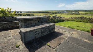 A stone bench with a stone wall backdrop overlooking a green field with buildings in the distance. - Pake's Cottage in Clahane (Liscannor)