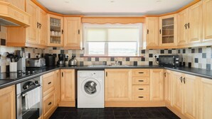A compact kitchen with wooden cabinets, a built-in washing machine, a microwave, and a window with blinds. - Pake's Cottage in Clahane (Liscannor)