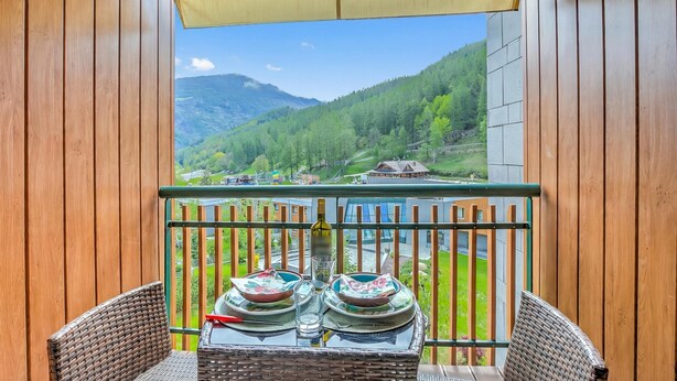 A balcony with a view of a mountainous landscape, wicker furniture, and a table set with plates and a bottle. - Il Mughetto Ski in / ski out (Bardonecchia)