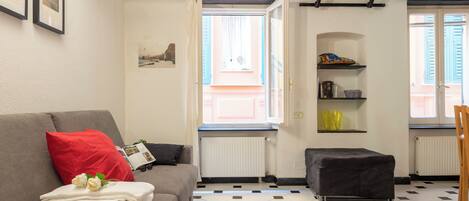 A living room with a grey sofa, a red pillow, a small black ottoman, and a tiled floor.